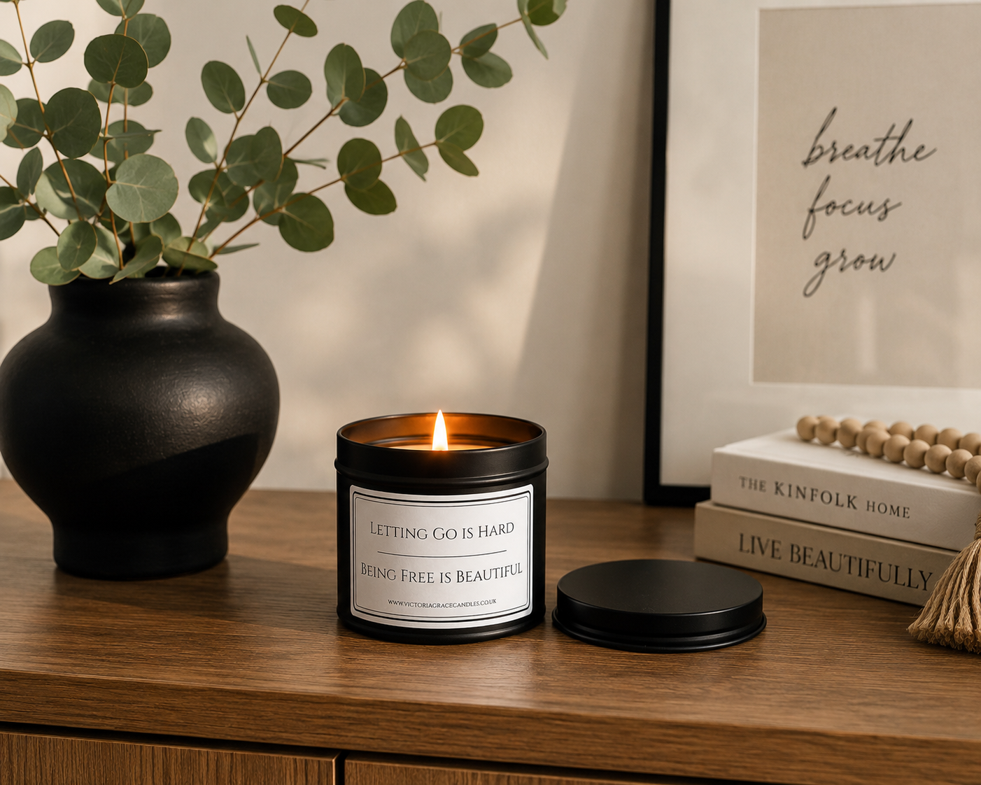 A lit black metal candle tin with a white label sits on a wooden table beside a black vase with eucalyptus and a framed wall print. The label reads, 'LETTING GO IS HARD being free is beautiful
