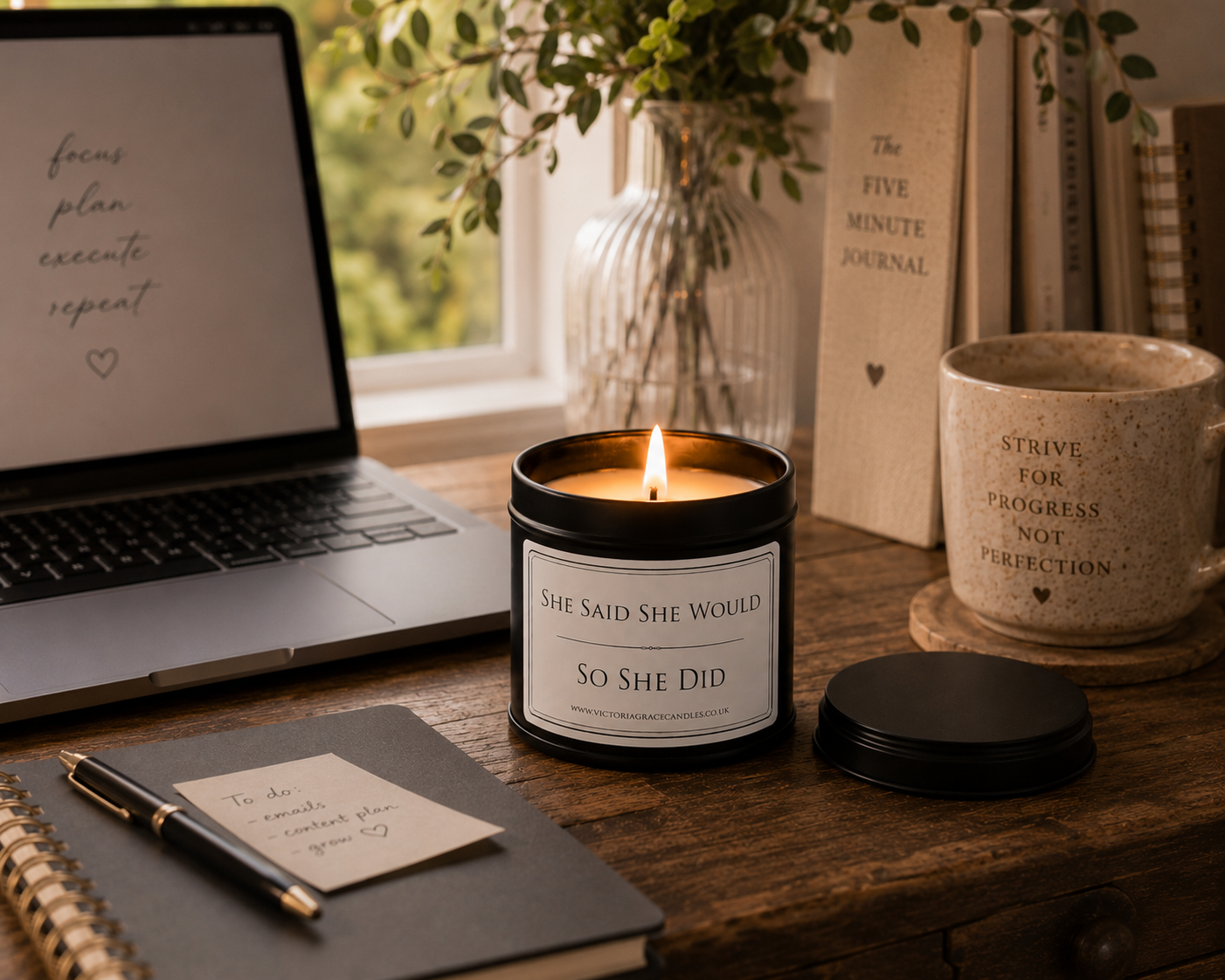 A lit black candle jar with a cream label reading 'SHE SAID SHE WOULD So SHE Did' sits on a wooden desk beside a laptop, notebook, pen, mug, books, and a glass vase with greenery in a bright indoor setting.