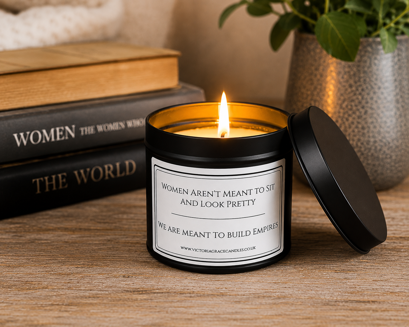 A black metal candle jar with a white label reading 'WOMEN AREN'T MEANT TO SIT AND LOOK PRETTY WE ARE MEANT TO BUILD EMPIRES' sits lit on a wooden table with its lid leaning beside it. Books and a potted plant are blurred in the background.
