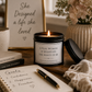 A black candle jar with a lit flame sits on a wooden table in a cozy styled setting, with a matching black lid beside it. The candle label reads, 'A REAL WOMAN IS WHATEVER SHE WANTS TO BE.
