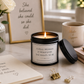 A black candle jar with a white label reading 'A REAL WOMAN IS WHATEVER SHE WANTS TO BE' sits on a desk beside a matching candle lid, notebook, flowers, mug, and framed sign in a bright home office setting.