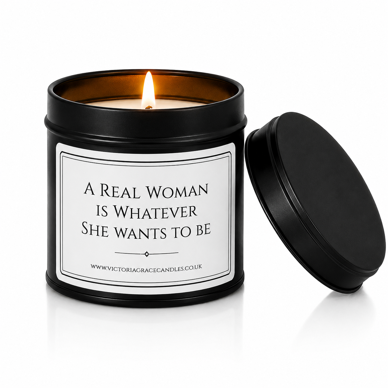 A black metal candle tin with a lit candle inside, shown next to its matching black lid on a white background. The front label reads, 'A REAL WOMAN IS WHATEVER SHE WANTS TO BE' 