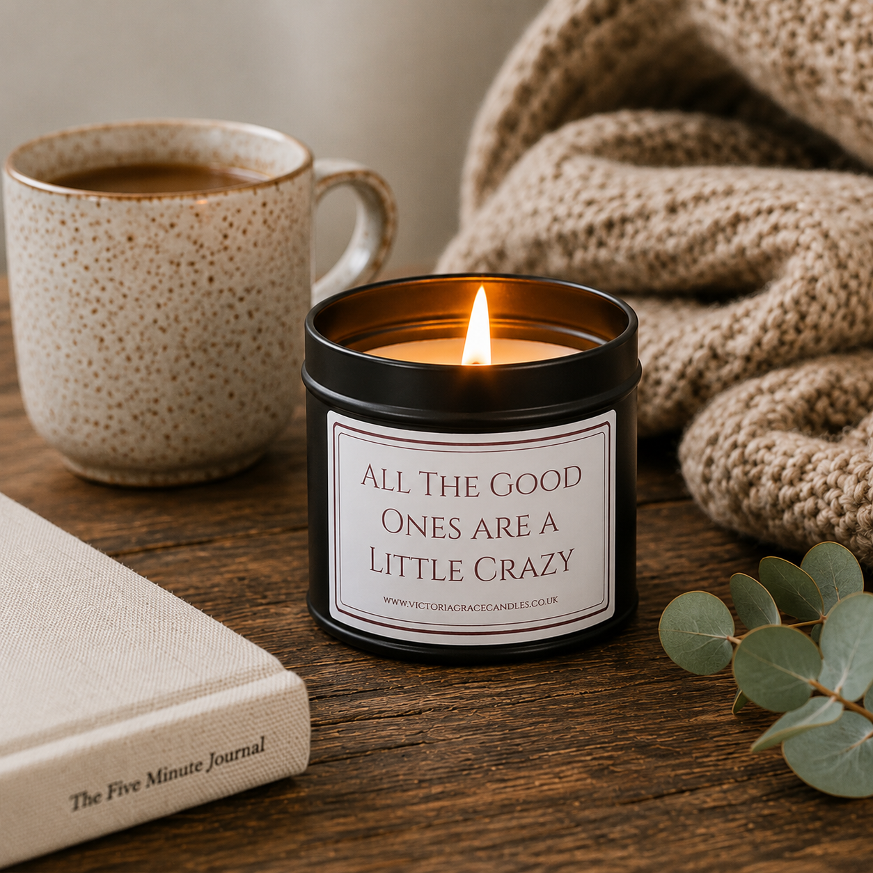 Luxury soy wax candle displayed on a rustic wooden coffee table beside a mug of coffee, cosy knitted blanket, journal and eucalyptus.