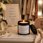 A lit candle in a black metal tin sits on a vanity table in a warm, styled room setting. The candle has a white label that reads, 'BE WITH THE GUY THAT RUMS YOUR LIPSTICK NOT YOUR MASCARA.