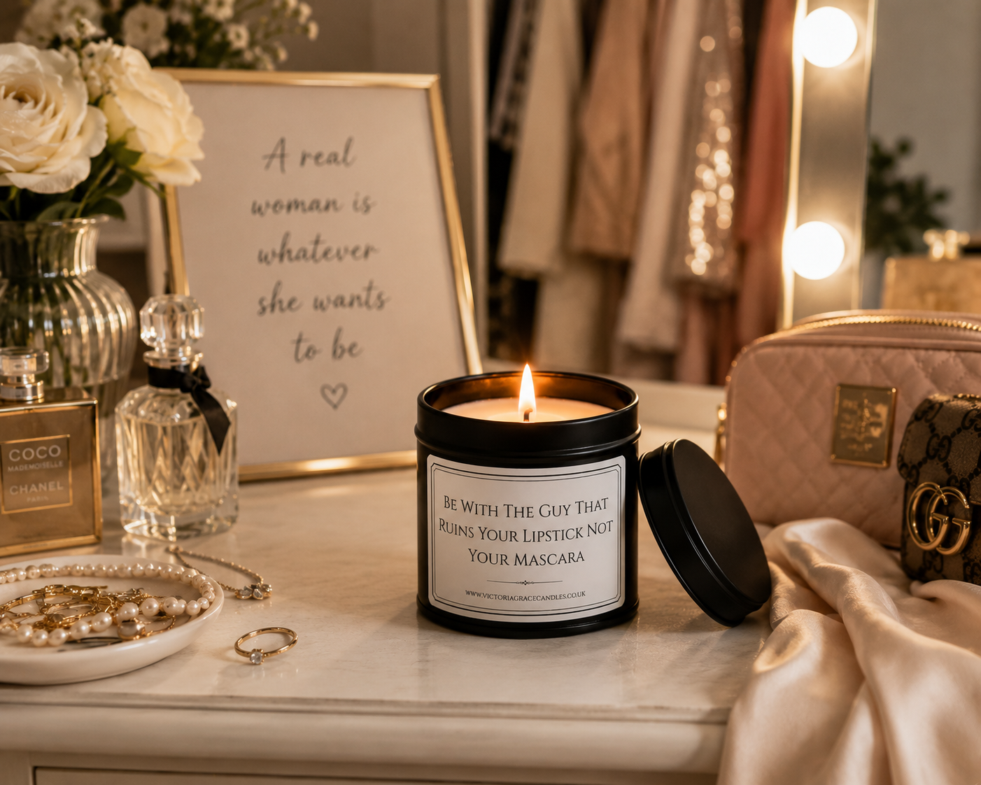 A lit candle in a black metal tin sits on a vanity table in a warm, styled room setting. The candle has a white label that reads, 'BE WITH THE GUY THAT RUMS YOUR LIPSTICK NOT YOUR MASCARA.