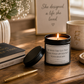 A lit black candle jar with a white label reading 'Be With The Guy That Being Your Lipstick Not Your Mascara' sits on a wooden table beside a black lid, a cream mug with gold lettering, a book, a pen, and a framed quote.