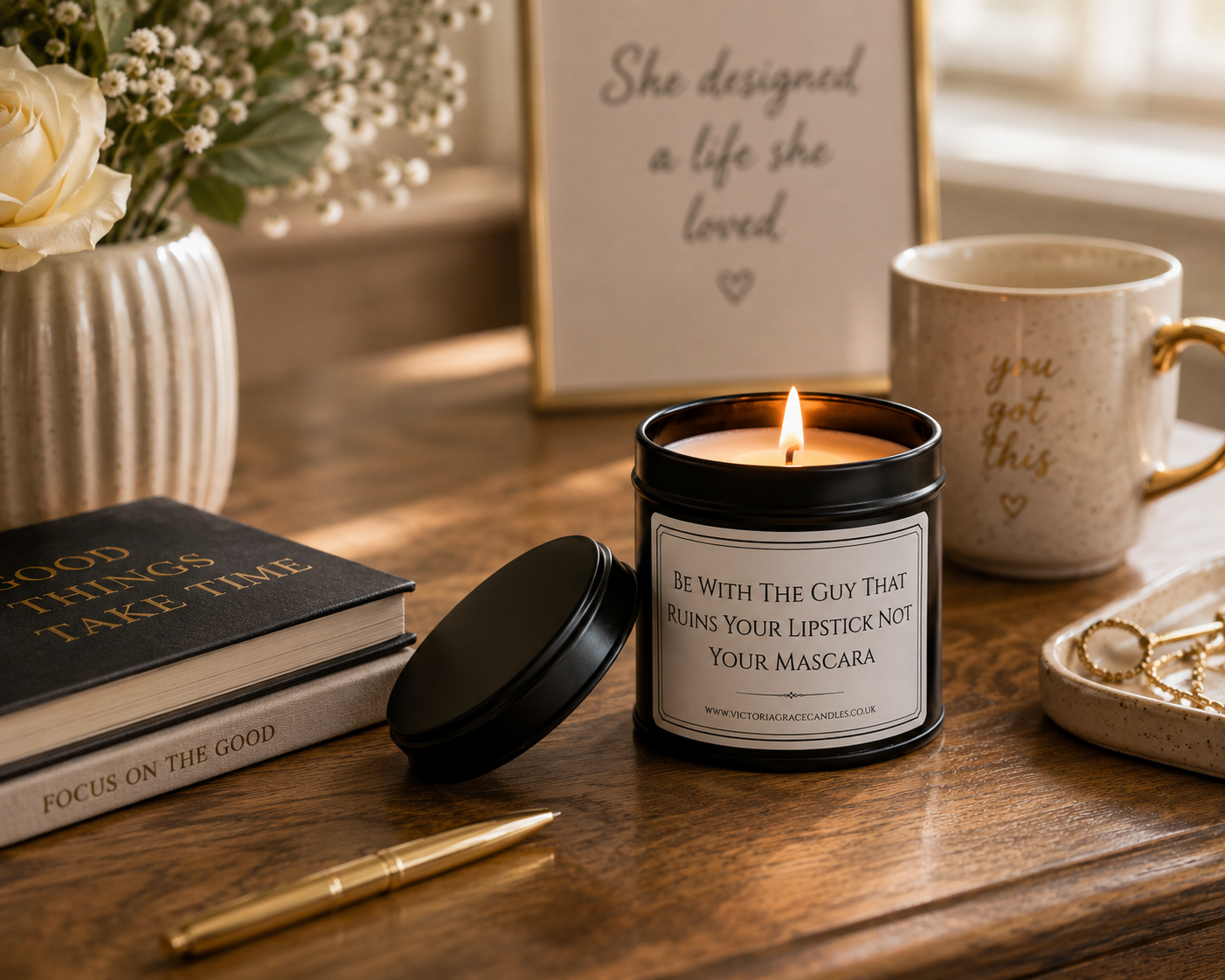 A lit black candle jar with a white label reading 'Be With The Guy That Being Your Lipstick Not Your Mascara' sits on a wooden table beside a black lid, a cream mug with gold lettering, a book, a pen, and a framed quote.