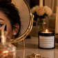 A lit candle in a black jar sits on a vanity table beside a round mirror showing part of a woman's face. The candle label reads, 'BE WITH THE GUY THAT RUNS YOUR LIPSTICK NOT YOUR MASCARA.