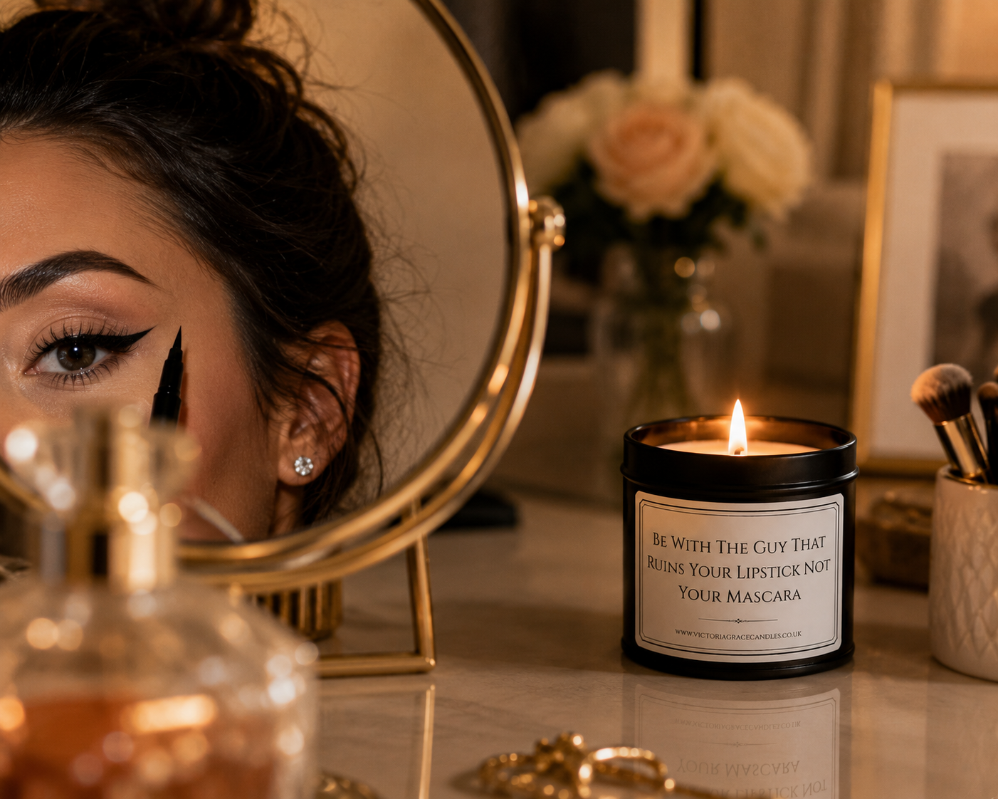 A lit candle in a black jar sits on a vanity table beside a round mirror showing part of a woman's face. The candle label reads, 'BE WITH THE GUY THAT RUNS YOUR LIPSTICK NOT YOUR MASCARA.