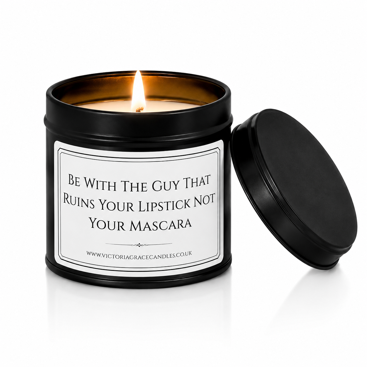 A black metal candle tin with a lit flame and a white printed label that reads, 'BE WITH THE GUY THAT RUINS YOUR LIPSTICK NOT YOUR MASCARA.' The lid rests beside the tin on a clean white background.