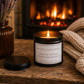 Luxury inspirational candle beside a glowing fireplace with a chunky knitted blanket and amber glass vase, creating a warm and peaceful setting.