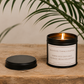 Luxury inspirational soy wax candle displayed on a rustic wooden table beneath tropical green leaves, creating a calm and uplifting atmosphere.