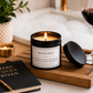 A lit black candle jar labeled BITCHCRAFT sits on a wooden tray beside its lid, with a wine glass, notebook, candle, and plant in a bathroom-like setting.