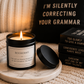 A lit black candle jar with a white label reading BITCHCRAFT sits on a round marble coaster, with the lid placed beside it. The scene is styled on a beige knit blanket with a black pillow that says “I’M SILENTLY CORRECTING YOUR GRAMMAR” and a stack of black books.