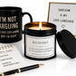 A curated product arrangement on a white background featuring a black candle tin with a lit flame and black lid, a black mug with gold text, a small notepad, a pen, and a framed sign reading 'SARCASM IS MY LOVE LANGUAGE'.