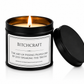 A black metal candle tin with a lit candle inside, shown on a white background with the lid leaning against it. The front label reads 'BITCHCRAFT' with a slogan below.