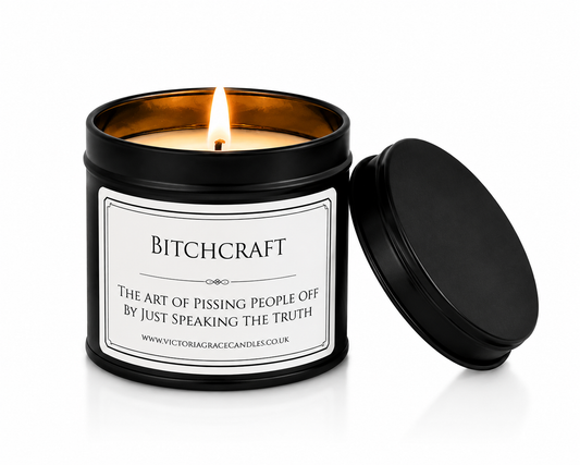 A black metal candle tin with a lit candle inside, shown on a white background with the lid leaning against it. The front label reads 'BITCHCRAFT' with a slogan below.