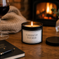 A lit candle in a black metal tin sits on a wooden table with its lid beside it. The label reads “CHEER UP FUCKER. background have a lit fireplace