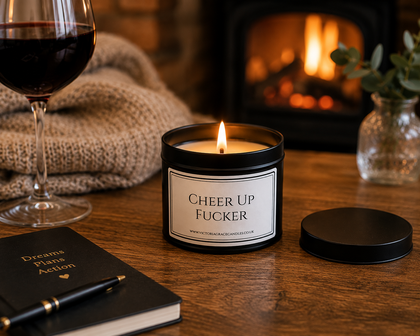A lit candle in a black metal tin sits on a wooden table with its lid beside it. The label reads “CHEER UP FUCKER. background have a lit fireplace