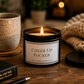 A black candle jar with a white label reading 'CHEER UP FUCKER' sits on a wooden table, lit with a warm flame. A speckled beige mug, a chunky knit blanket, and a black book are nearby in a cozy indoor setting.