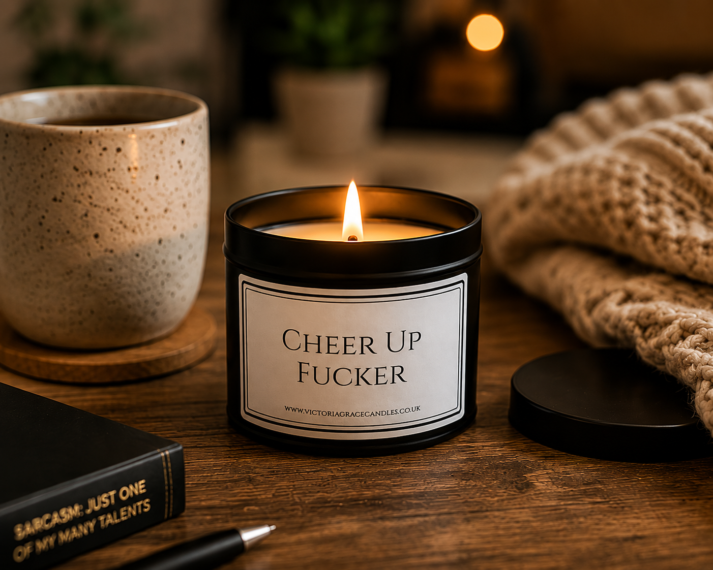 A black candle jar with a white label reading 'CHEER UP FUCKER' sits on a wooden table, lit with a warm flame. A speckled beige mug, a chunky knit blanket, and a black book are nearby in a cozy indoor setting.
