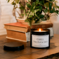 Luxury black soy wax candle displayed on a wooden table with books and greenery in a cosy home setting.