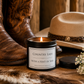 A lit candle in a black tin labeled 'Country Lass with a Hint of Sass' sits on a rustic wooden surface with cowboy boots, a straw hat, and small white flowers around it.