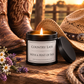 A lit black metal candle tin labeled 'COUNTRY LASS WITH A HINT OF SASS' sits on a rustic wooden surface outdoors at sunset, surrounded by cowboy boots, a straw hat, flowers, and rope.