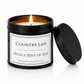 A lit scented candle in a matte black metal tin with its lid resting beside it on a white background. The label reads 'COUNTRY LASS WITH A HINT OF SASS' and shows the brand website 