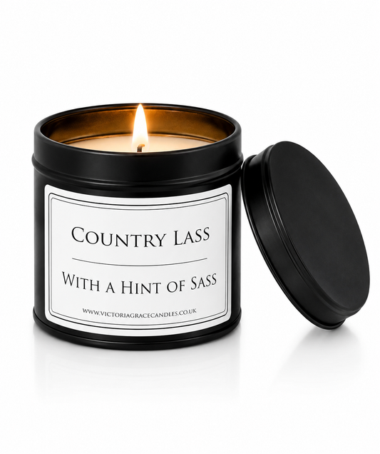 A lit scented candle in a matte black metal tin with its lid resting beside it on a white background. The label reads 'COUNTRY LASS WITH A HINT OF SASS' and shows the brand website 