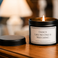 A black metal candle jar with a lit flame sits on a wooden table next to its lid. The front label reads, 'Dance Like No One Is Watching' with the brand 