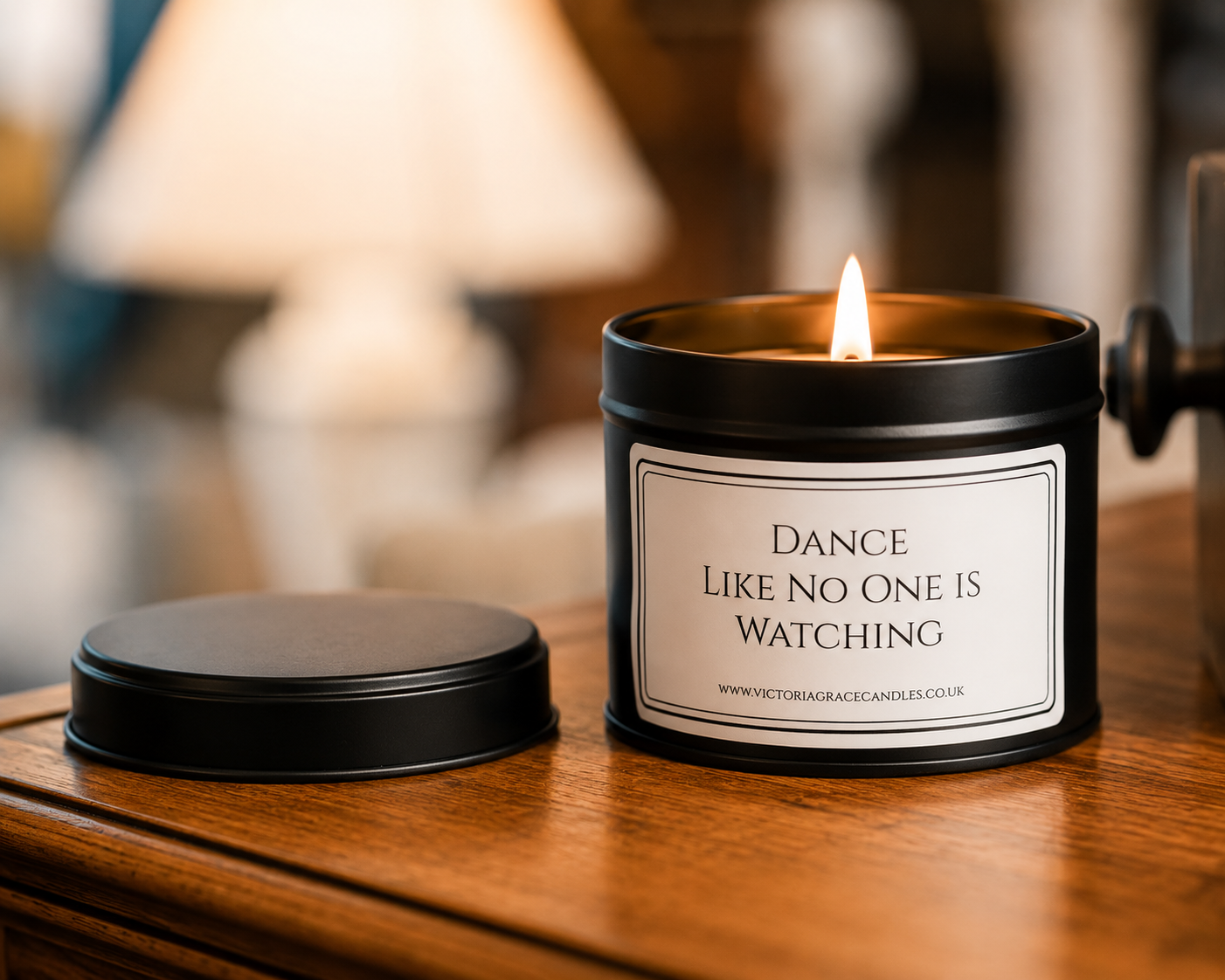 A black metal candle jar with a lit flame sits on a wooden table next to its lid. The front label reads, 'Dance Like No One Is Watching' with the brand 