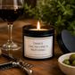 A lit candle in a black tin jar sits on a wooden table with a wine glass, corkscrew, olives, and greenery in the background. The front label reads, "Dance Like No One Is Watching.