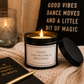 A black metal candle jar with a white label reading 'Dance Like No One Is Watching' sits on a cream knit blanket, with its lid beside it. A lit candle in a glass holder, a black notebook with gold text, and a black pillow with gold text are arranged around it in a cozy indoor setting.