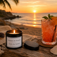 Luxury black soy wax candle beside a tropical cocktail overlooking a sandy beach and the sea at sunset, creating the perfect holiday setting.