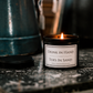 A lit black jar candle labeled 'Drink In Hand / Toes In Sand' sits on a dark marble surface with its lid beside it. A teal metal container is blurred in the background.