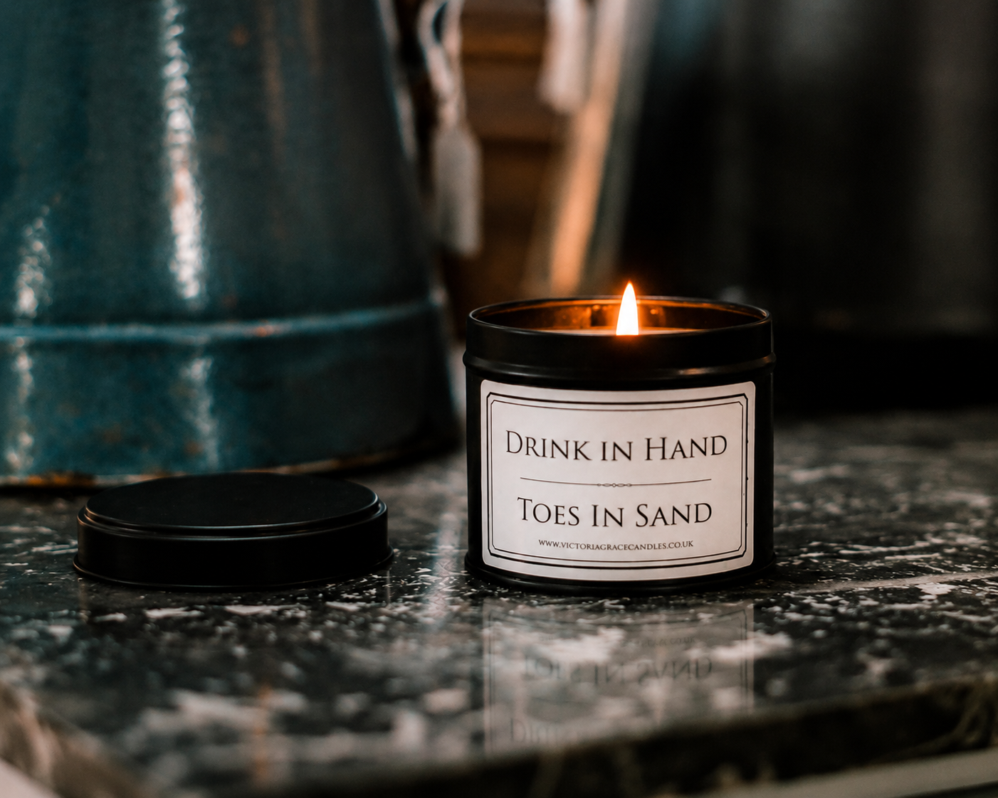 A lit black jar candle labeled 'Drink In Hand / Toes In Sand' sits on a dark marble surface with its lid beside it. A teal metal container is blurred in the background.