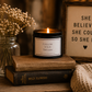 A lit black candle jar with a white label reading 'FOLLOW YOUR DREAMS' sits on stacked vintage books beside a glass jar of dried flowers and a framed sign that says 'SHE BELIEVED SHE COULD SO SHE DID'.