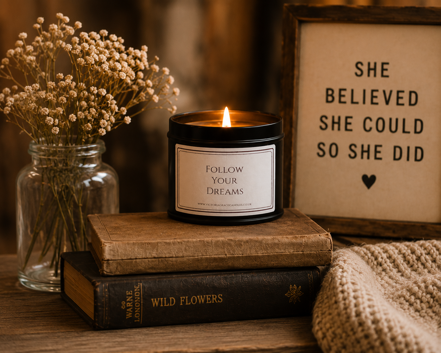 A lit black candle jar with a white label reading 'FOLLOW YOUR DREAMS' sits on stacked vintage books beside a glass jar of dried flowers and a framed sign that says 'SHE BELIEVED SHE COULD SO SHE DID'.