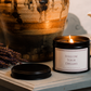 A lit black metal candle jar with a cream label reading 'FOLLOW YOUR DREAMS' sits on a tabletop beside its lid, styled with dried lavender and a large ceramic vase in the background.