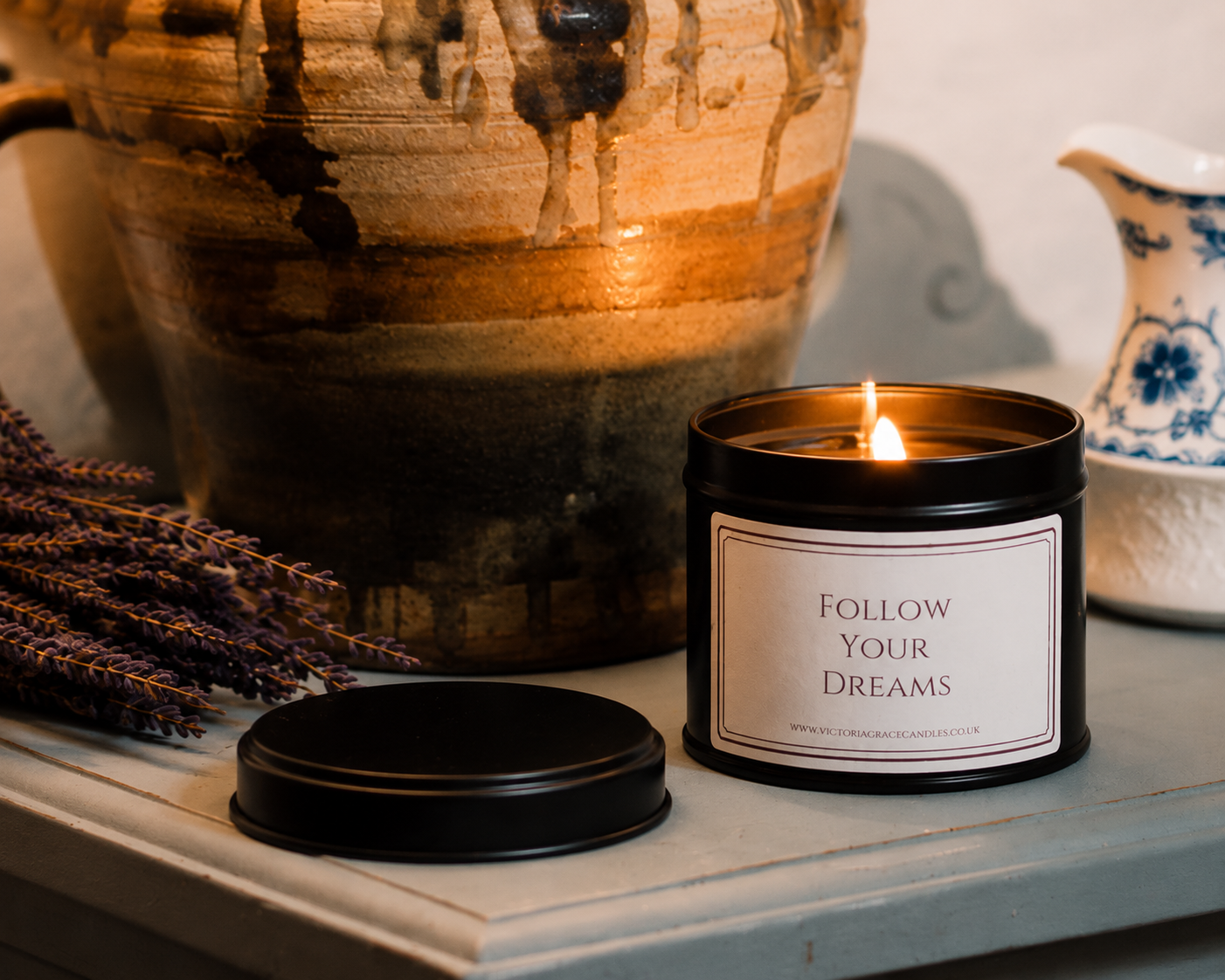A lit black metal candle jar with a cream label reading 'FOLLOW YOUR DREAMS' sits on a tabletop beside its lid, styled with dried lavender and a large ceramic vase in the background.