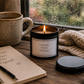 A black metal candle tin with a white label reading 'FOLLOW YOUR DREAMS' sits lit on a wooden windowsill beside a ceramic mug, a notebook with handwritten text, a pen, and a knitted beige scarf.