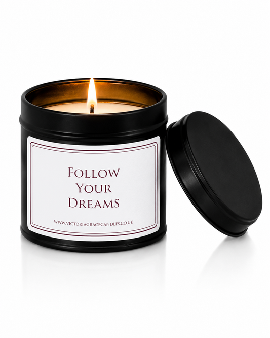 A black metal candle tin with a lit wick and flame, featuring a white label that reads 'FOLLOW YOUR DREAMS' and the website 