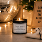 A black metal candle jar with a white label reading 'FRIDAY NIGHTS ARE FOR KITCHEN DISCOS' sits on a tabletop with a lit candle inside. The scene is warmly lit with string lights, a disco ball, a folded striped towel, and a sign that says 'DANCE LIKE NO ONE'S WASHING UP'.