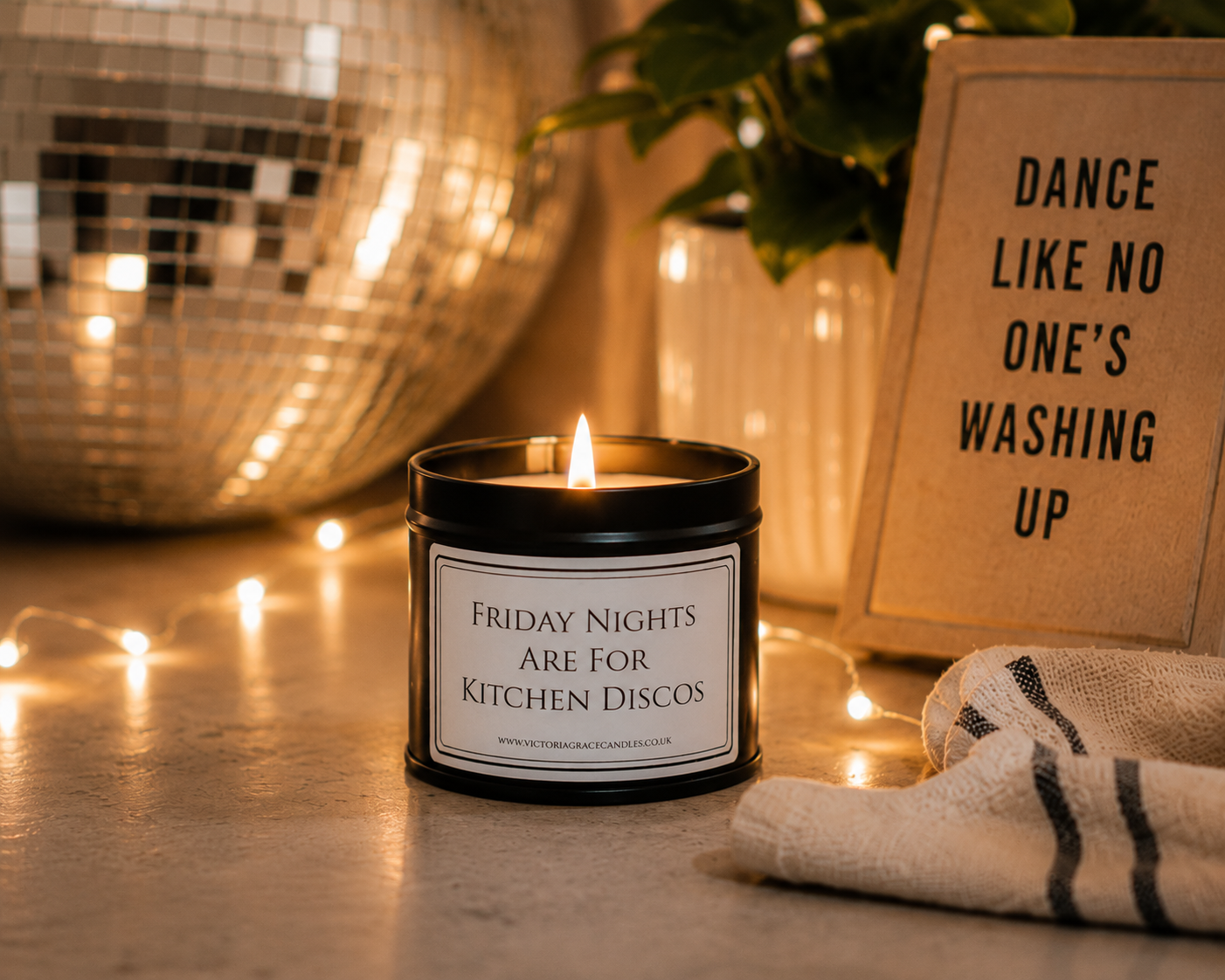 A black metal candle jar with a white label reading 'FRIDAY NIGHTS ARE FOR KITCHEN DISCOS' sits on a tabletop with a lit candle inside. The scene is warmly lit with string lights, a disco ball, a folded striped towel, and a sign that says 'DANCE LIKE NO ONE'S WASHING UP'.