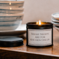 A black metal candle tin with a white label reading 'FRIDAY NIGHTS ARE FOR KITCHEN DISCOS' sits lit on a wooden surface, with its lid placed beside it and stacked ceramic bowls blurred in the background.