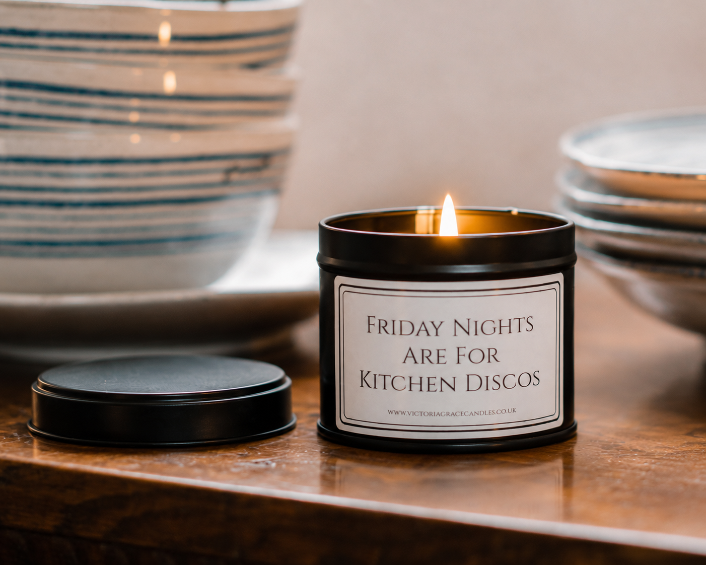 A black metal candle tin with a white label reading 'FRIDAY NIGHTS ARE FOR KITCHEN DISCOS' sits lit on a wooden surface, with its lid placed beside it and stacked ceramic bowls blurred in the background.