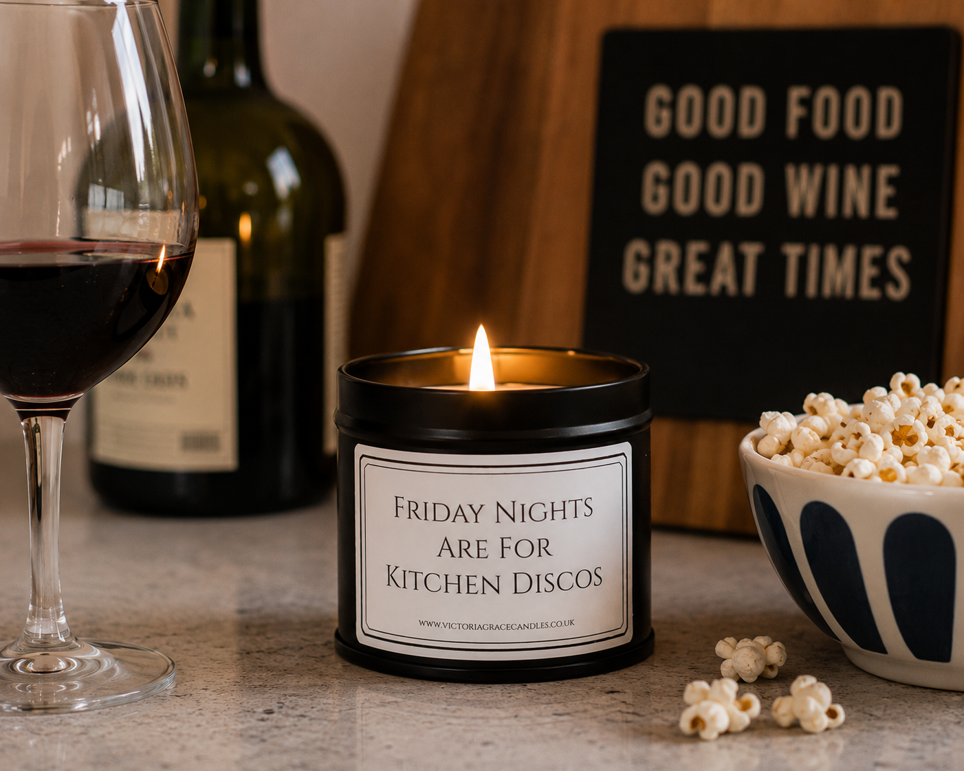A black candle tin with a lit flame sits on a countertop, surrounded by a wine glass, a bottle, popcorn, and a sign that reads 'GOOD FOOD GOOD WINE GREAT TIMES'. The candle label says 'FRIDAY NIGHTS ARE FOR KITCHEN DISCOS'.
