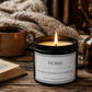 A lit black candle jar with a white label reading 'HOME It's not a place it's a feeling' sits on a rustic wooden table with a mug, book, and dried flowers in the background.