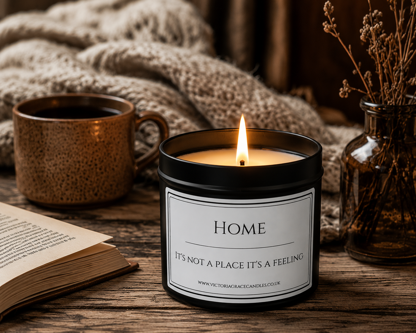 A lit black candle jar with a white label reading 'HOME It's not a place it's a feeling' sits on a rustic wooden table with a mug, book, and dried flowers in the background.